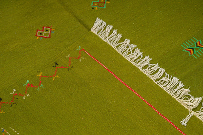 A close-up of a green moroccan rug handwoven with geometric embroidered motifs in red, yellow, and teal, plus a prominent white fringe running diagonally across the surface.