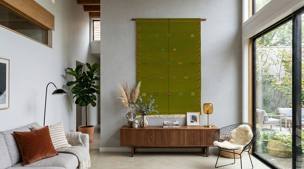 A green woven moroccan wall hanging rug or tapestry with small embroidered motifs hangs on a white wall in a modern, sunlit living room. A wooden sideboard, sofa, chair, and large windows are visible in the room.