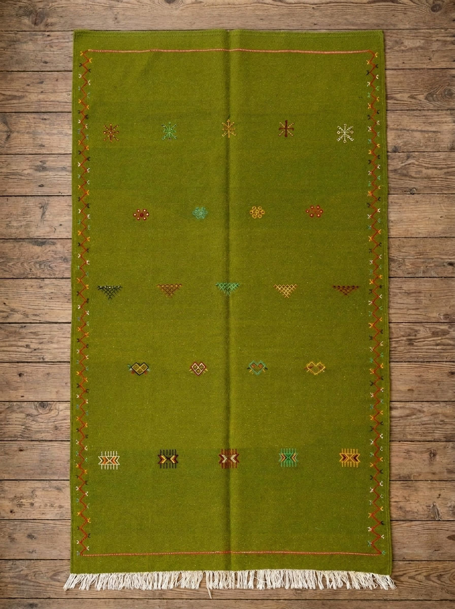 An olive green moroccan wool rug with white fringe at the bottom and small embroidered multicolor geometric patterns, laid flat on a rustic wooden plank floor.
