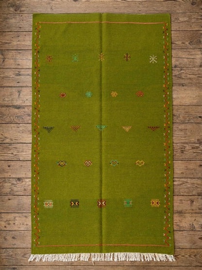 An olive green moroccan wool rug with white fringe at the bottom and small embroidered multicolor geometric patterns, laid flat on a rustic wooden plank floor.