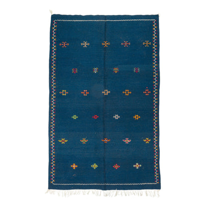 A rectangular blue moroccan rug with a hand embroidered geometric pattern of small colorful diamond motifs and white fringe at the top and bottom, shown flat against a white background.