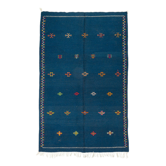 A rectangular blue moroccan rug with a hand embroidered geometric pattern of small colorful diamond motifs and white fringe at the top and bottom, shown flat against a white background.