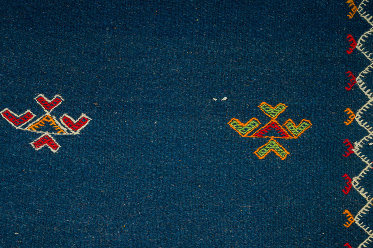 Close-up of a dark blue moroccan rug from akhnif with small geometric embroidered motifs in red, yellow, and green, plus a decorative border along the right edge.