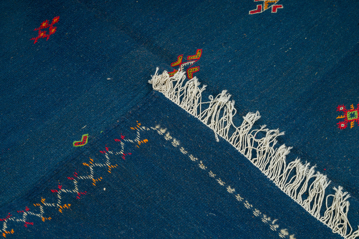 A close-up of a blue moroccan rug , featuring a prominent white fringe trim and small multicolored embroidered geometric motifs.