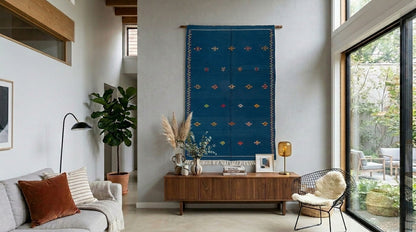 A blue moroccan wall hanging rug with embroidered geometric patterns is displayed in a modern, bright living room above a wooden console table.