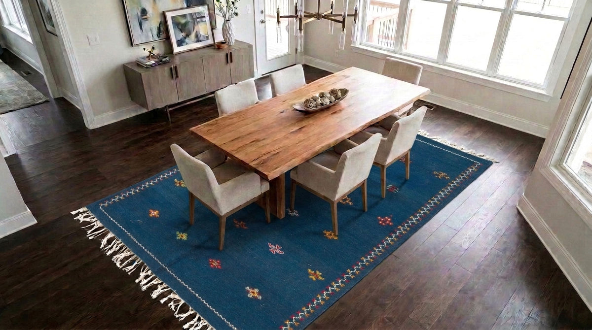 A blue moroccan rug with embroidered motifs placed under a rectangular wooden dining table with six upholstered chairs in a bright dining room with dark wood floors and large windows.
