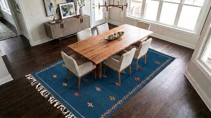 A blue moroccan rug with embroidered motifs placed under a rectangular wooden dining table with six upholstered chairs in a bright dining room with dark wood floors and large windows.
