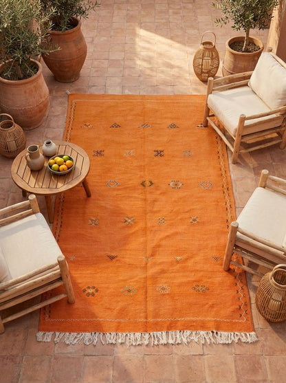An orange Moroccan handwoven area rug with cream fringe and small geometric motifs, styled on a terracotta tile patio surrounded by wooden chairs, potted plants, and a small wooden side table.