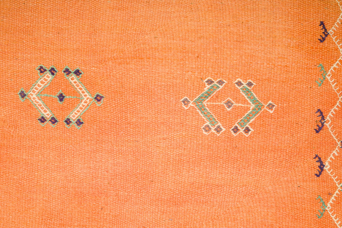 Close-up of an orange moroccan rug with embroidered geometric motifs showing the wool weave texture and part of a patterned border on the right edge.