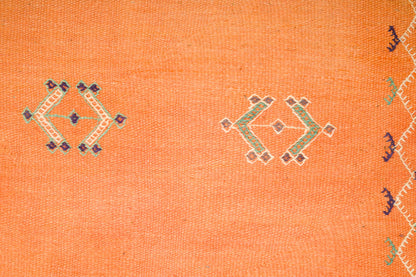 Close-up of an orange moroccan rug with embroidered geometric motifs showing the wool weave texture and part of a patterned border on the right edge.