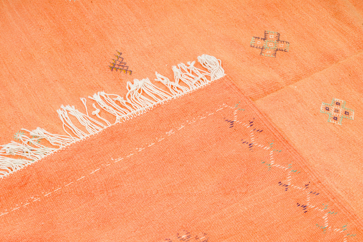 A close-up of a peach-orange moroccan rug with white fringe along one edge and small embroidered geometric motifs in muted multicolor thread.