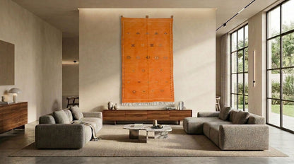 A modern living room featuring a large moroccan orange wall hanging rug artwork centered above a wooden console table, flanked by two gray upholstered sofas and a low round coffee table. Large windows on the right show greenery outside, and the room has warm neutral walls and a rug.