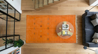 An overhead view of a bright orange Moroccan area rug with hand embroidered geometric motifs and fringe, styled in a modern living room with a gray sofa, wooden staircase, and light wood floors.
