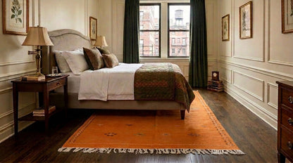 Moroccan orange rug set over dark wood flooring in a styled bedroom interior featuring a bed with white bedding and a patterned green throw. The room has cream paneled walls, dark green curtains, and a window looking out onto a city scene.