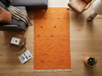 An orange rectangular area rug with white fringe and small geometric motifs, laid out on a light wood floor in a styled living room setting with a gray sofa, leather chair, side table, and plant.