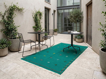 A rectangular green moroccan rug with a subtle floral pattern and fringe, styled on a light stone patio next to a black metal dining table and chairs.