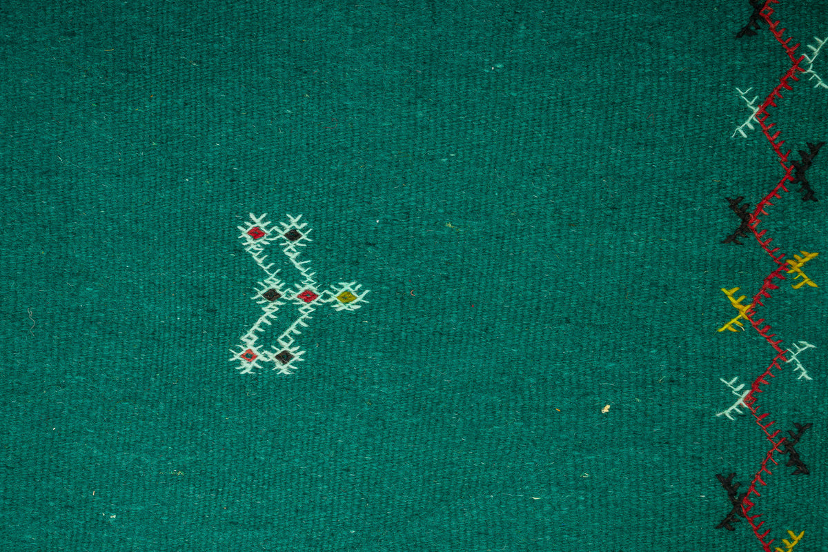 Close-up of a green moroccan rug with embroidered geometric and zigzag patterns in white, red, yellow, and black.