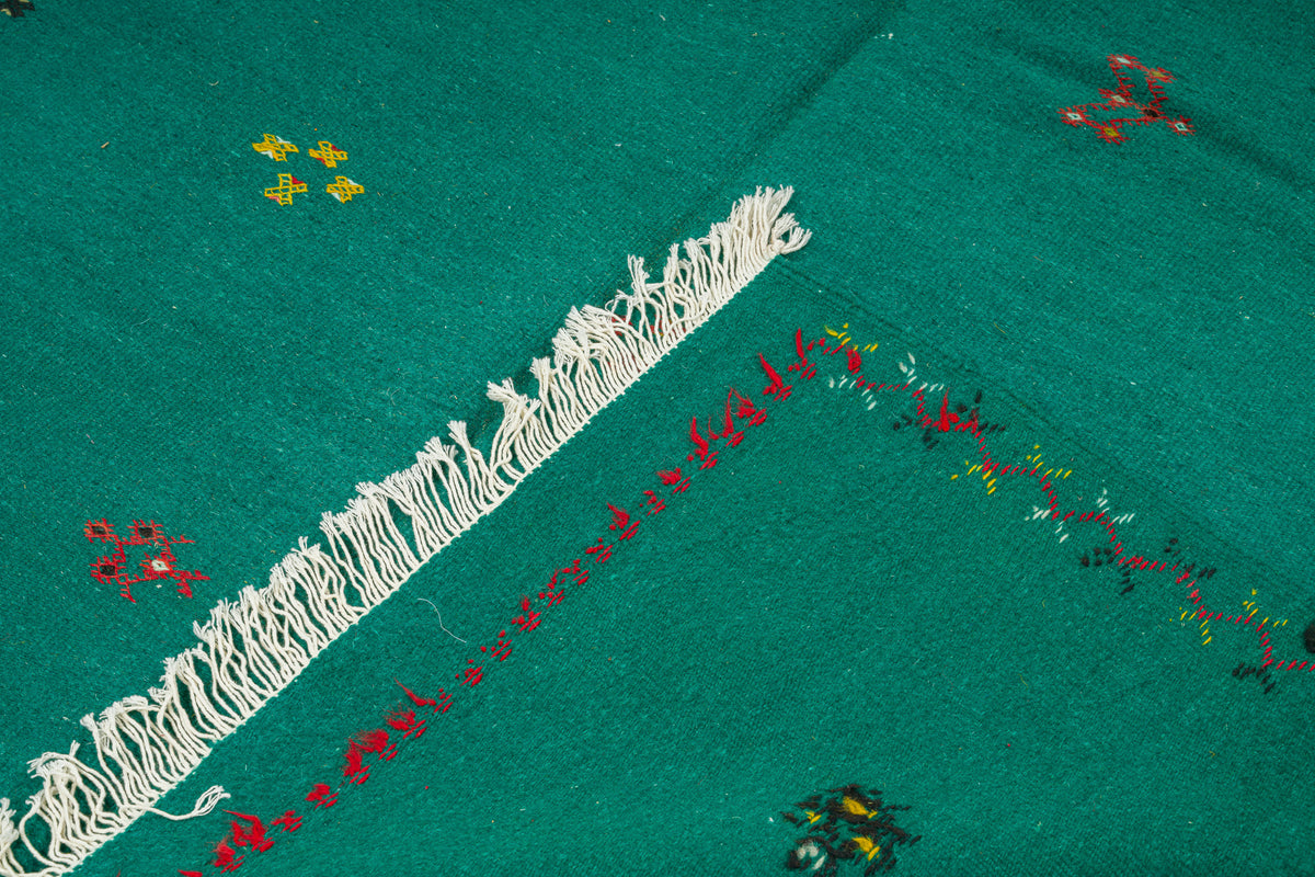 A close-up of a green moroccan rug, featuring a prominent white fringe trim, red embroidered border details, and small multicolored floral motifs.