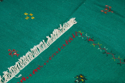 A close-up of a green moroccan rug, featuring a prominent white fringe trim, red embroidered border details, and small multicolored floral motifs.
