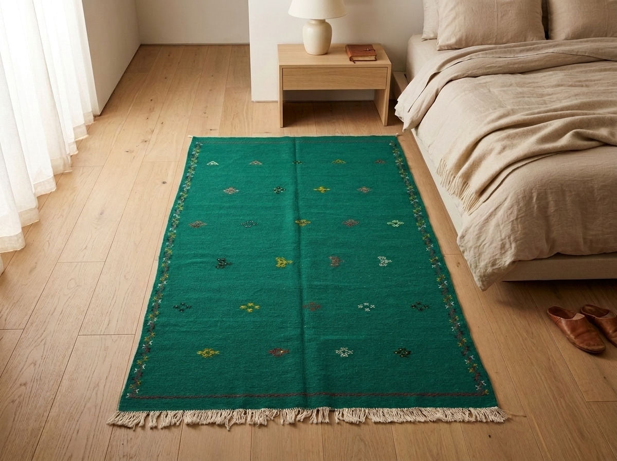 A green rectangular moroccan area rug with cream fringe and small embroidered multicolor geometric motifs, styled on a light wood floor next to a bed in a bedroom setting.