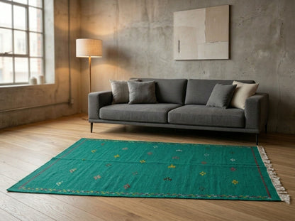 An emerald green moroccan area rug with small multicolored tribal motifs and white fringe, styled on a light wood floor in a modern loft living room with a gray sofa and concrete walls.