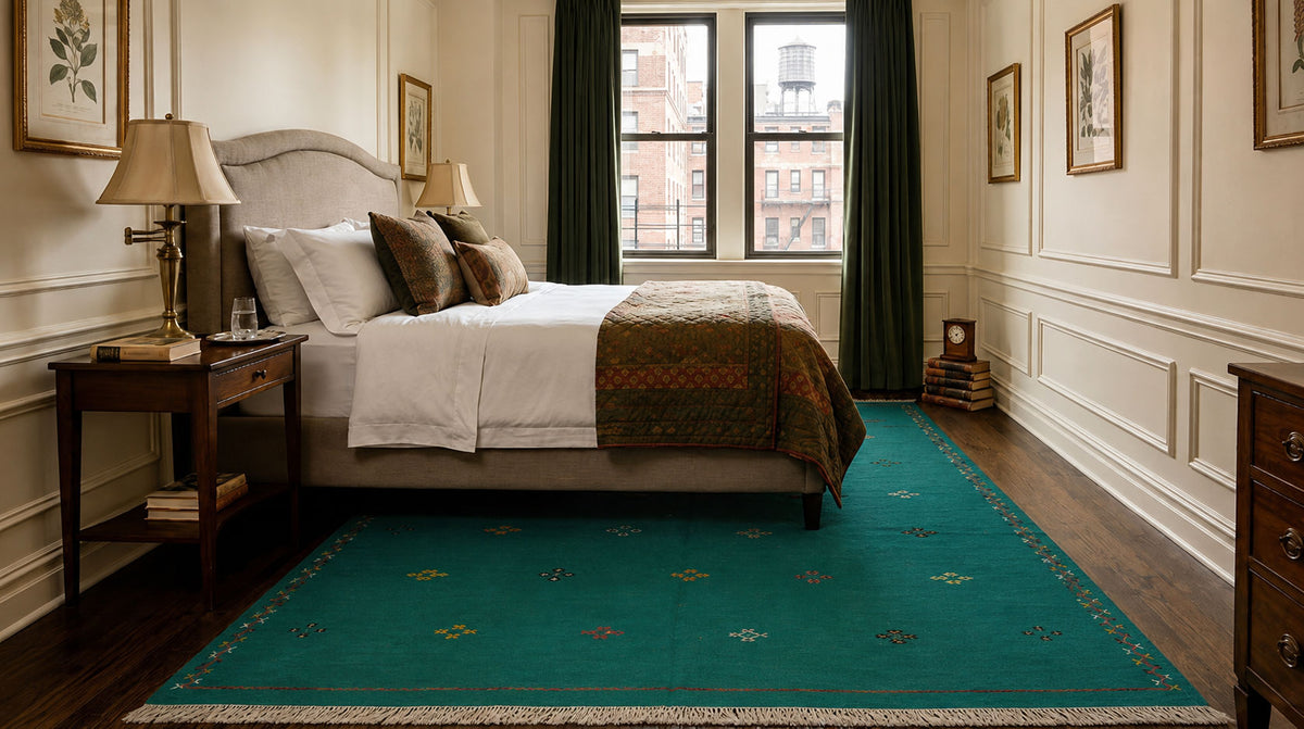 A green moroccan area rug with a subtle geometric pattern and cream fringe, styled in a classic bedroom setting with a bed, nightstand, and window.