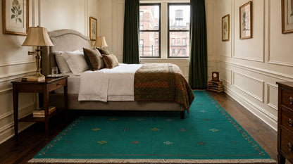 A green moroccan area rug with a subtle geometric pattern and cream fringe, styled in a classic bedroom setting with a bed, nightstand, and window.