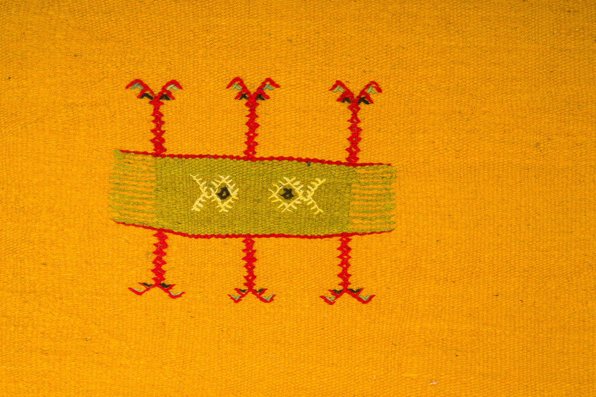A close-up of a yellow moroccan rug featuring a central rectangular hand embroidered motif in olive green with red embroidered legs and small yellow details.