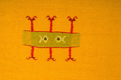 A close-up of a yellow moroccan rug featuring a central rectangular hand embroidered motif in olive green with red embroidered legs and small yellow details.
