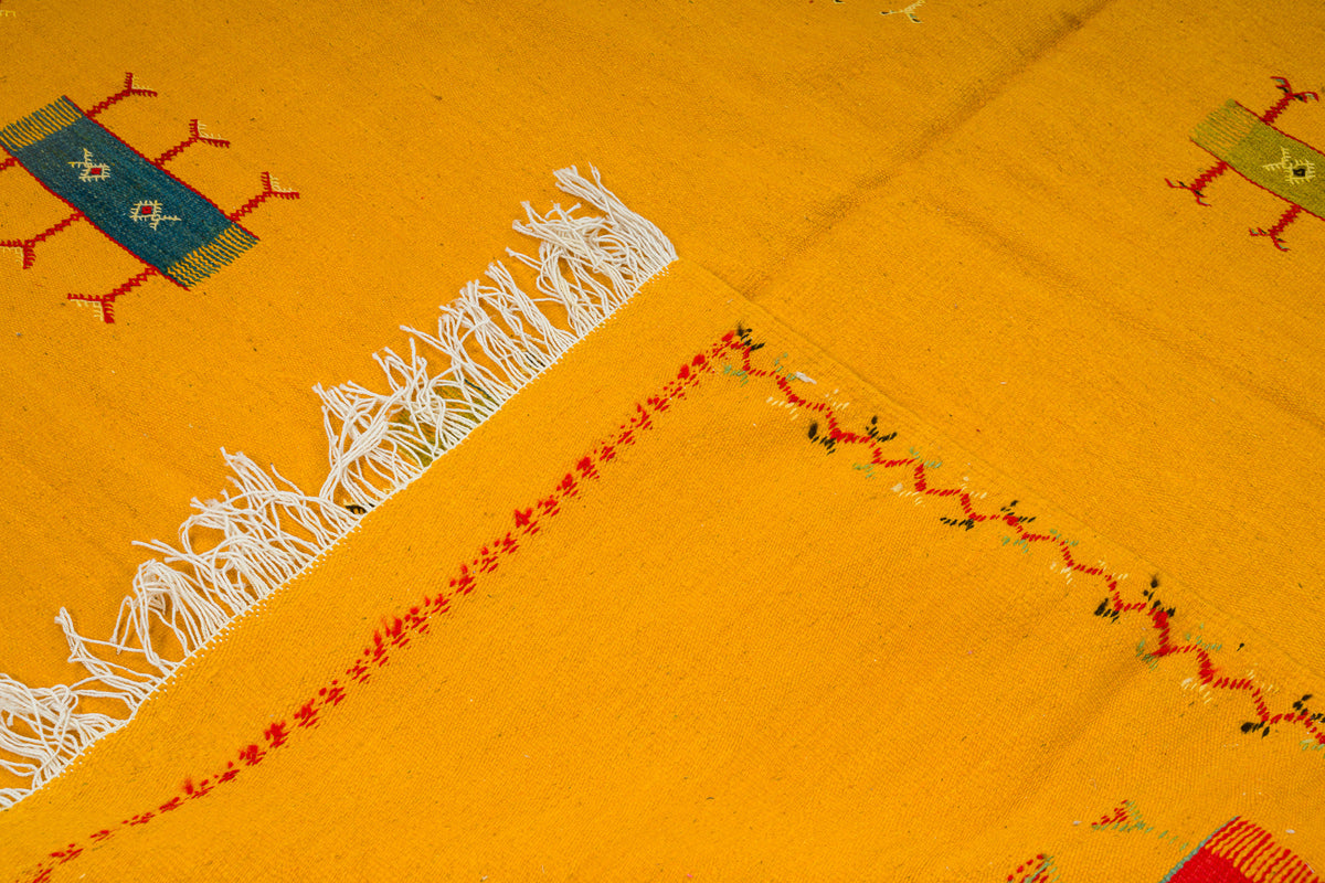 A close-up of a mustard yellow handwoven moroccan rug, featuring white fringe, red and black embroidered lines, and small geometric motifs in blue, green, and red.