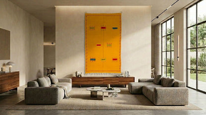 A modern living room featuring a large, vibrant yellow Moroccan tapestry rug hanging on the wall above a wooden console. The room is styled with two gray sectional sofas, a low wooden coffee table, and large floor-to-ceiling windows on the right.