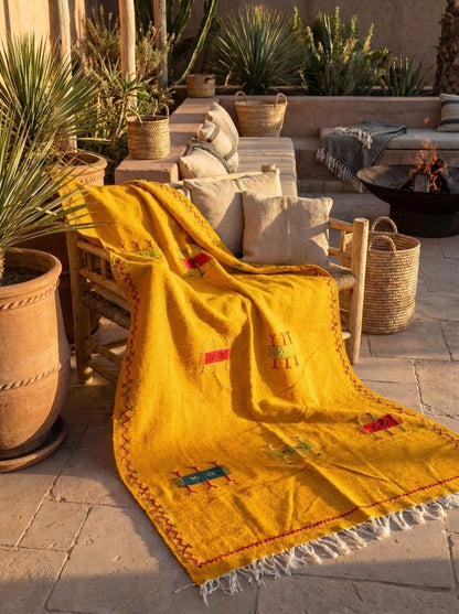 A bright yellow Moroccan rug with white fringe and colorful embroidered details is draped over a wooden lounge chair on a stone patio surrounded by potted plants.