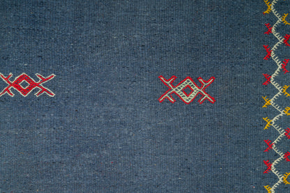Close-up of a moroccan blue rug with embroidered geometric motifs in red, white, and yellow along the right edge.