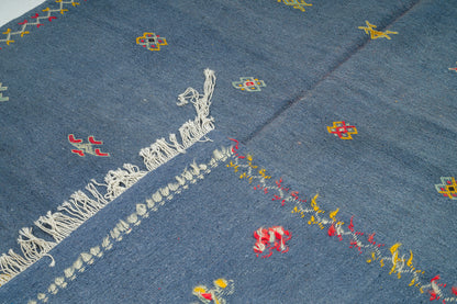 A close-up of a blue moroccan rug handwoven with white fringe on one side and scattered small geometric and floral motifs in red, yellow, and green.