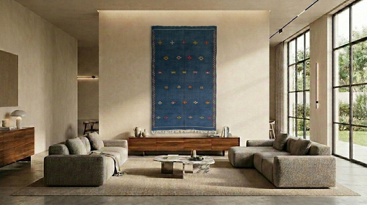 A large blue Moroccan handwoven wall hanging rug  with colorful geometric patterns is displayed in a modern, sunlit living room with gray sofas and a wooden console table.