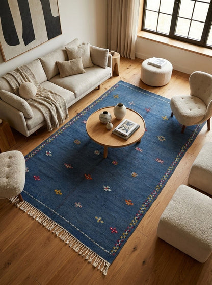 A blue area rug with cream fringe and small multicolor floral motifs is styled in a bright, modern living room with a beige sofa, wooden coffee table, and light wood floors.