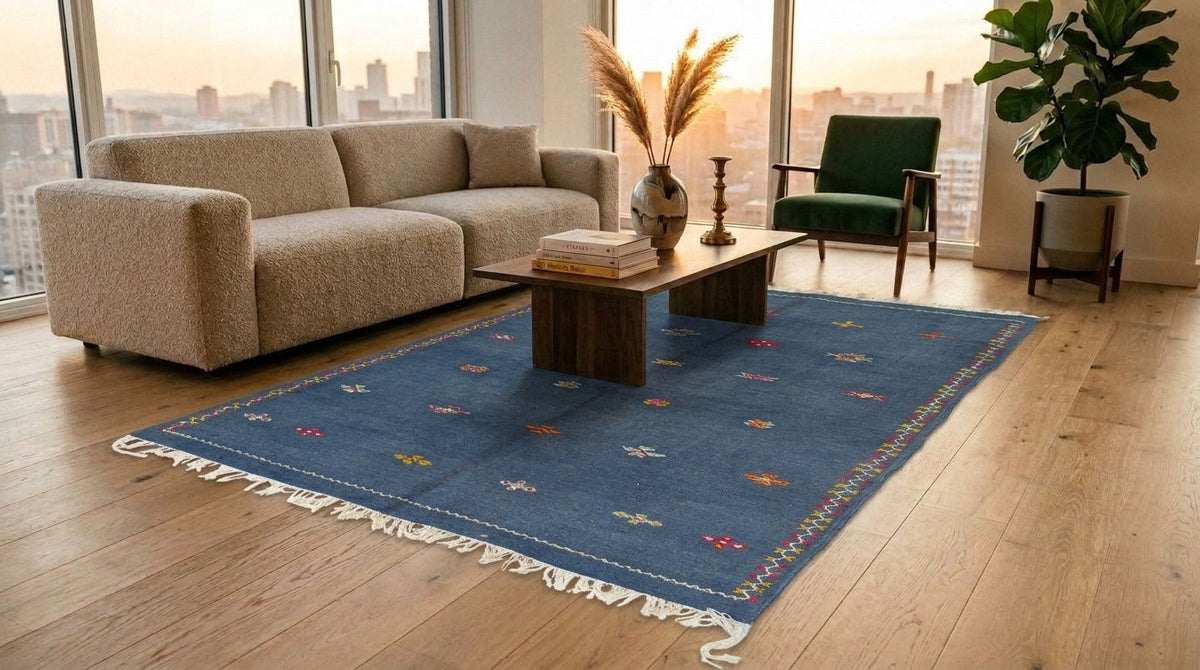 A blue rectangular moroccan area rug with white fringe and small multicolor embroidered motifs is styled in a modern living room with a beige sofa, wooden coffee table, green armchair, and large windows overlooking a city skyline at sunset.