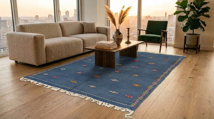 A blue rectangular moroccan area rug with white fringe and small multicolor embroidered motifs is styled in a modern living room with a beige sofa, wooden coffee table, green armchair, and large windows overlooking a city skyline at sunset.