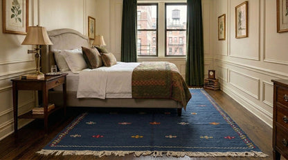 A styled moroccan blue area rug with fringe under a bed with white linens and a green patterned throw, dark wood nightstands, and a window with dark green curtains.