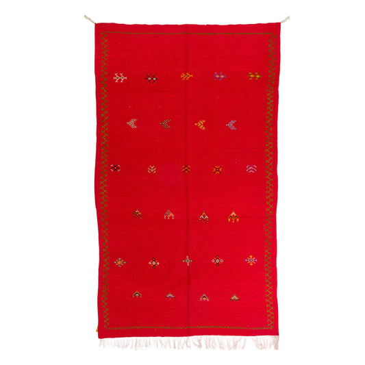 A red handwoven moroccan rug or scarf with a decorative border and small multicolored geometric motifs, shown flat against a white background.