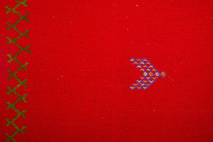 A close-up of a red moroccan rug with a vertical row of green embroidered crosses on the left and a small multicolor embroidered motif on the right.