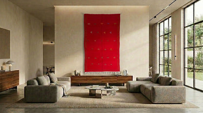 A modern living room featuring a large, vibrant red moroccan wall hanging tapestry centered above a wooden console table. The room is styled with two gray modular sofas, a low glass-top coffee table, and a textured beige rug, with large windows on the right showing a view of green trees.