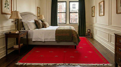 A red moroccan area rug with fringe  and hand embroidered motifs is styled in a classic bedroom setting with a bed, nightstand, and window in the background.
