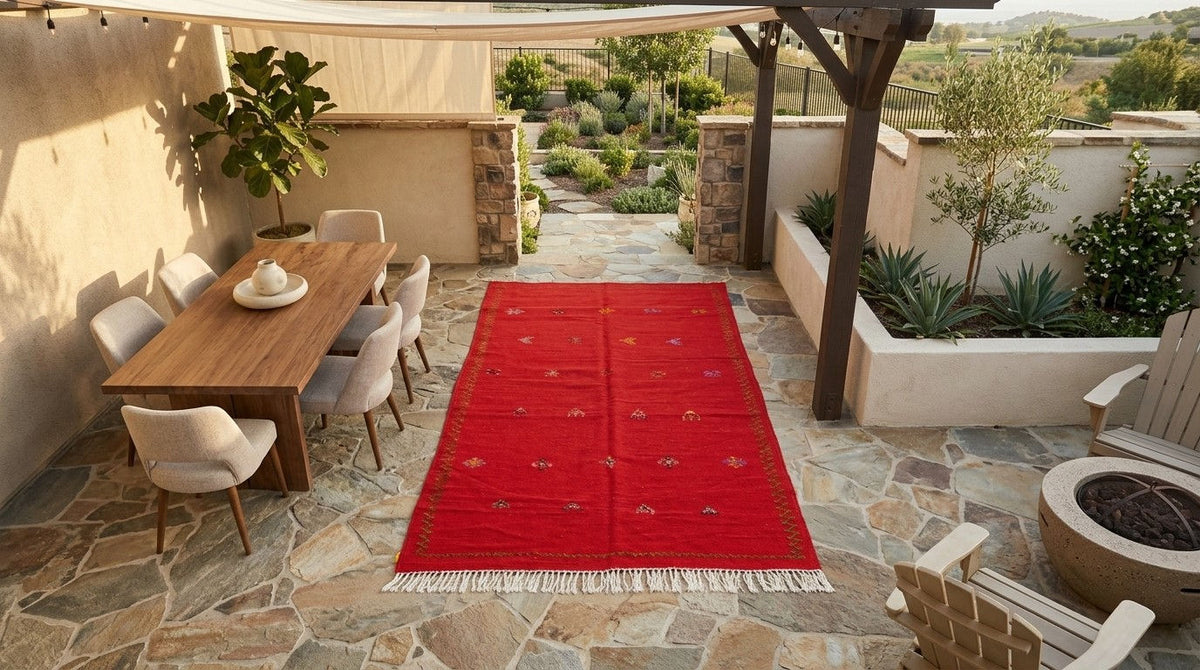 A vibrant red outdoor area rug with white fringe and small gold embroidered motifs, displayed on a stone patio in a modern backyard setting.