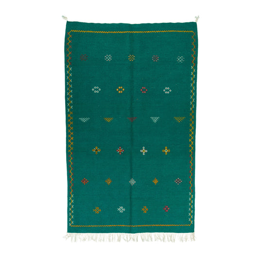 A green moroccan rug  handwoven with hand embroidered geometric pattern with small colorful diamond and cross motifs and white fringe at the bottom.