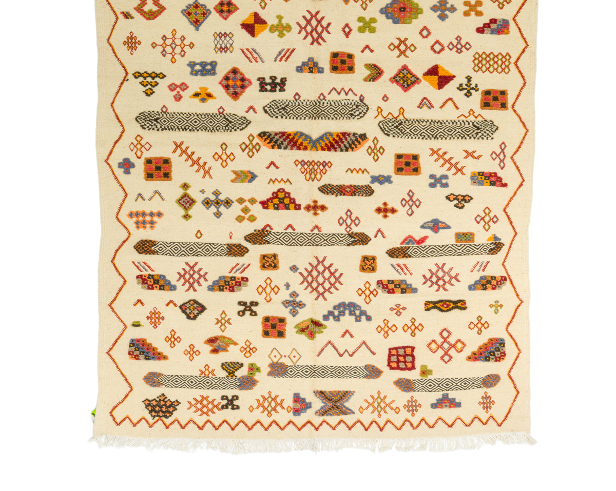 A flat-laid handwoven Moroccan rug with a cream-colored base and a dense, multicolored geometric pattern. The design features rows of stylized fish, zig-zag borders, and various diamond and cross motifs in red, orange, brown, and blue, finished with white fringe on the bottom edge.