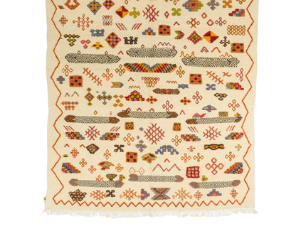 A flat-laid handwoven Moroccan rug with a cream-colored base and a dense, multicolored geometric pattern. The design features rows of stylized fish, zig-zag borders, and various diamond and cross motifs in red, orange, brown, and blue, finished with white fringe on the bottom edge.