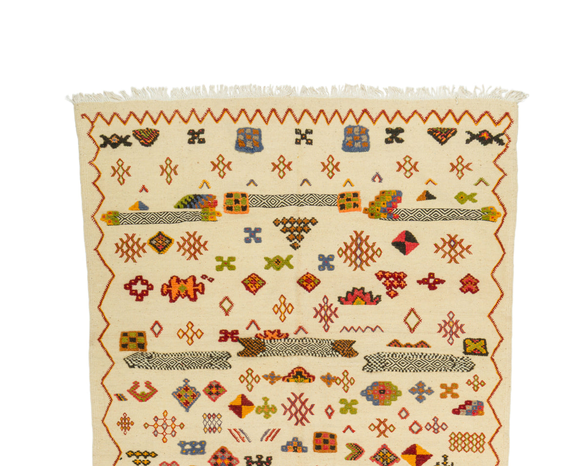 A square woven rug with a cream background and a colorful geometric pattern, featuring white fringe on the top edge, shown against a plain white background.