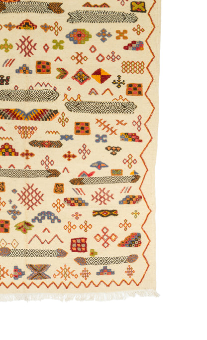 A close-up of a patterned rug with a cream background and multicolored geometric motifs, including orange, red, blue, green, and brown details, with a fringed edge visible at the bottom.