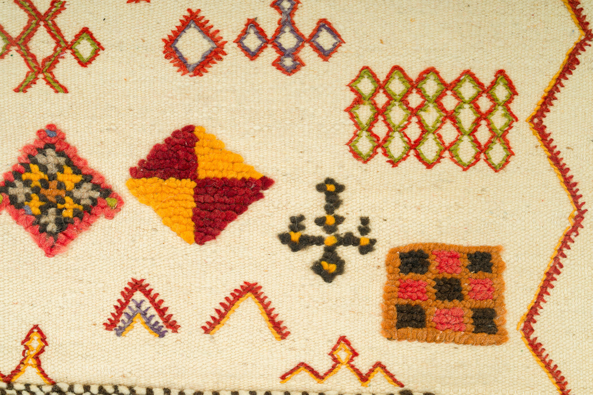 Close-up of a moroccan rug with geometric patterns in red, yellow, green, black, and pink on an off-white background.